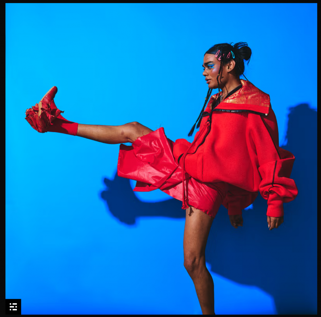 A person in bright red clothing performs a high kick against a vivid blue backdrop, casting a strong shadow that highlights their dynamic, energetic pose. 